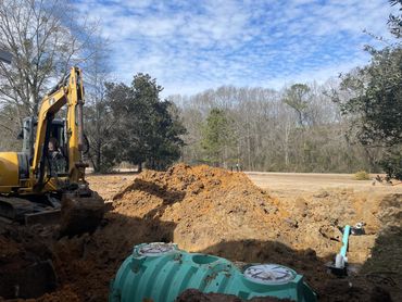 Excavator digging near a large underground tank with pipes in a rural area.