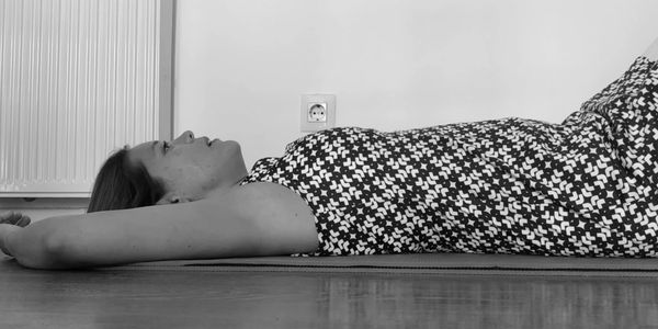 A woman lies on her back with arms extended, wearing a patterned dress indoors.