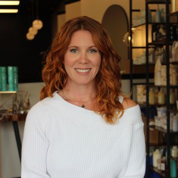 Smiling woman with red curly hair in a cozy white sweater inside a modern store.