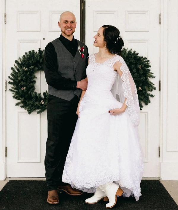 Couple in wedding attire with bride wearing white boots and groom in vest, standing in front of holiday wreaths.