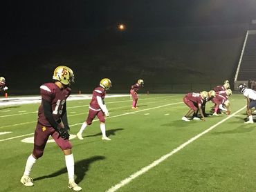 High school football players line up for a play under stadium lights at night.