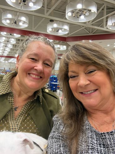 Two smiling women taking a selfie indoors under bright ceiling lights.