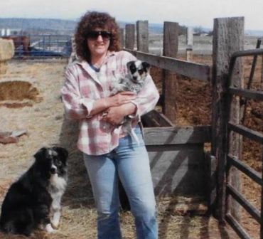 Woman in plaid shirt holding a puppy with another dog sitting nearby on a farm.