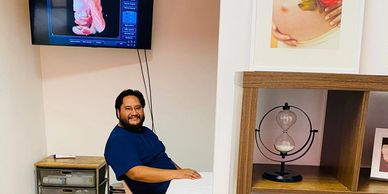 Man sitting in a medical office with a smile, an ultrasound image on screen behind him.