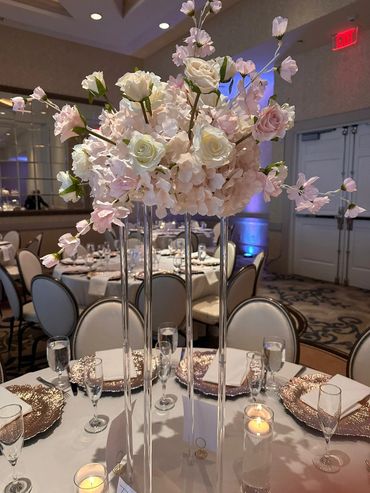 Elegant floral centerpiece with white and pink roses on tall clear stands at a banquet table.