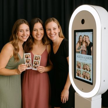 Three women smiling and holding photo booth strips beside a photo booth machine.