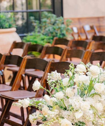 Elegant white floral arrangement beside wooden chairs at an outdoor event.