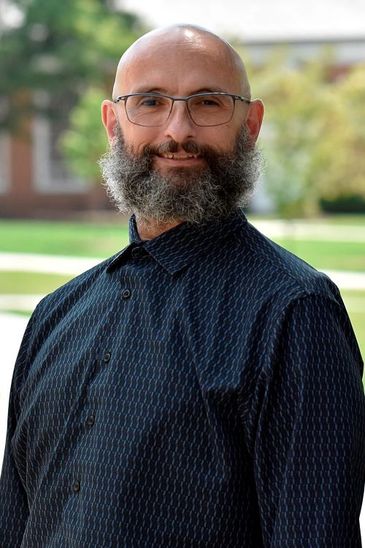 Smiling bald man with glasses and a full beard wearing a patterned dark shirt.
