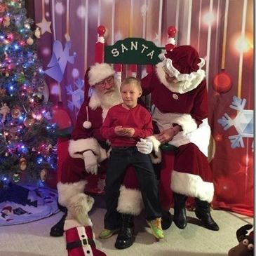 Santa and Mrs. Claus posing with child in Santa's chair with Christmas backdrop.