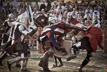 Medieval knights in armor engage in combat during a reenactment with an audience.