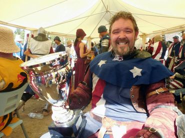 A man in medieval armor holding a large trophy and smiling under a tent.
