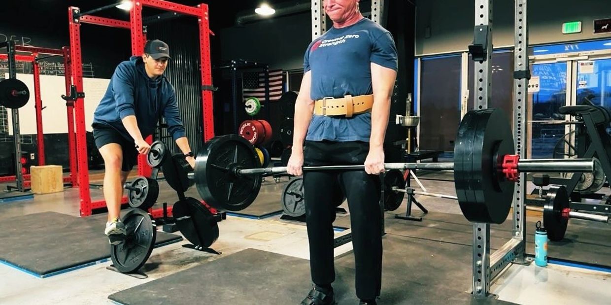 Man deadlifting heavy weights in a gym while another person watches.
