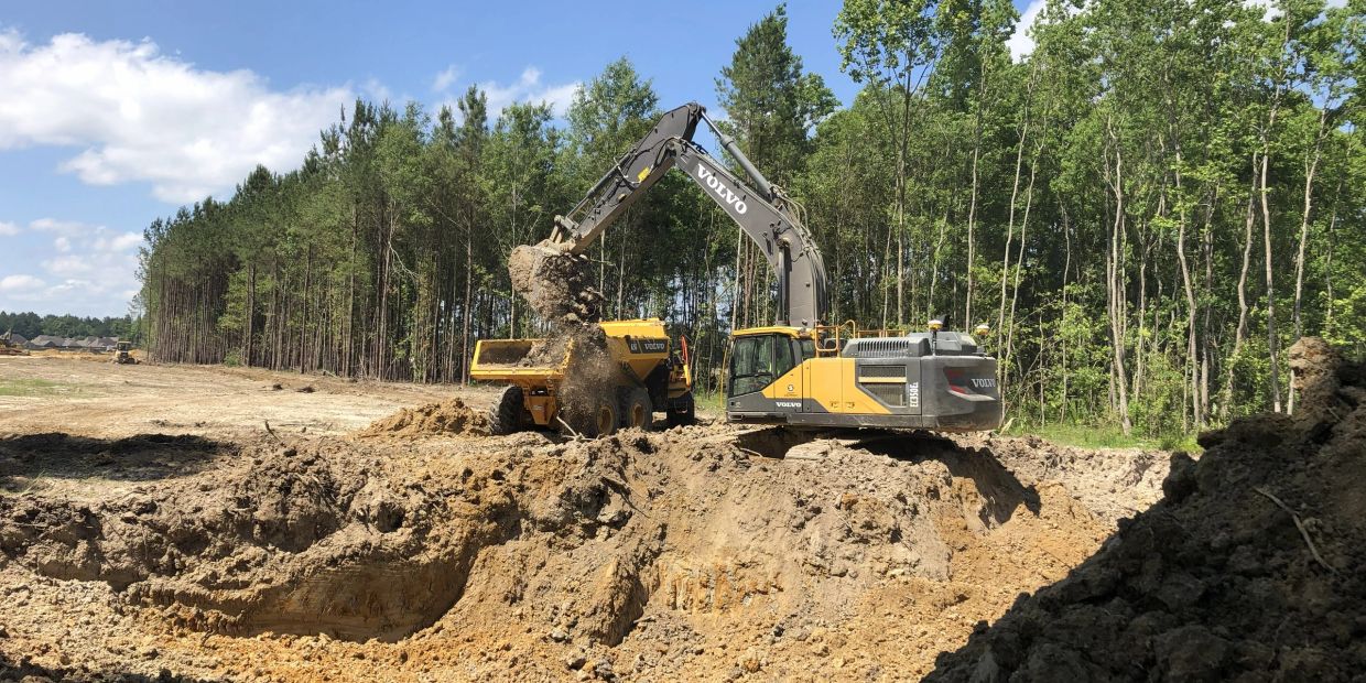 A Volvo excavator loading dirt into a dump truck at a construction site near a forest.