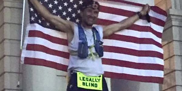 Jason in running gear in front of white pillars, smiling and raising an American flag behind him.
