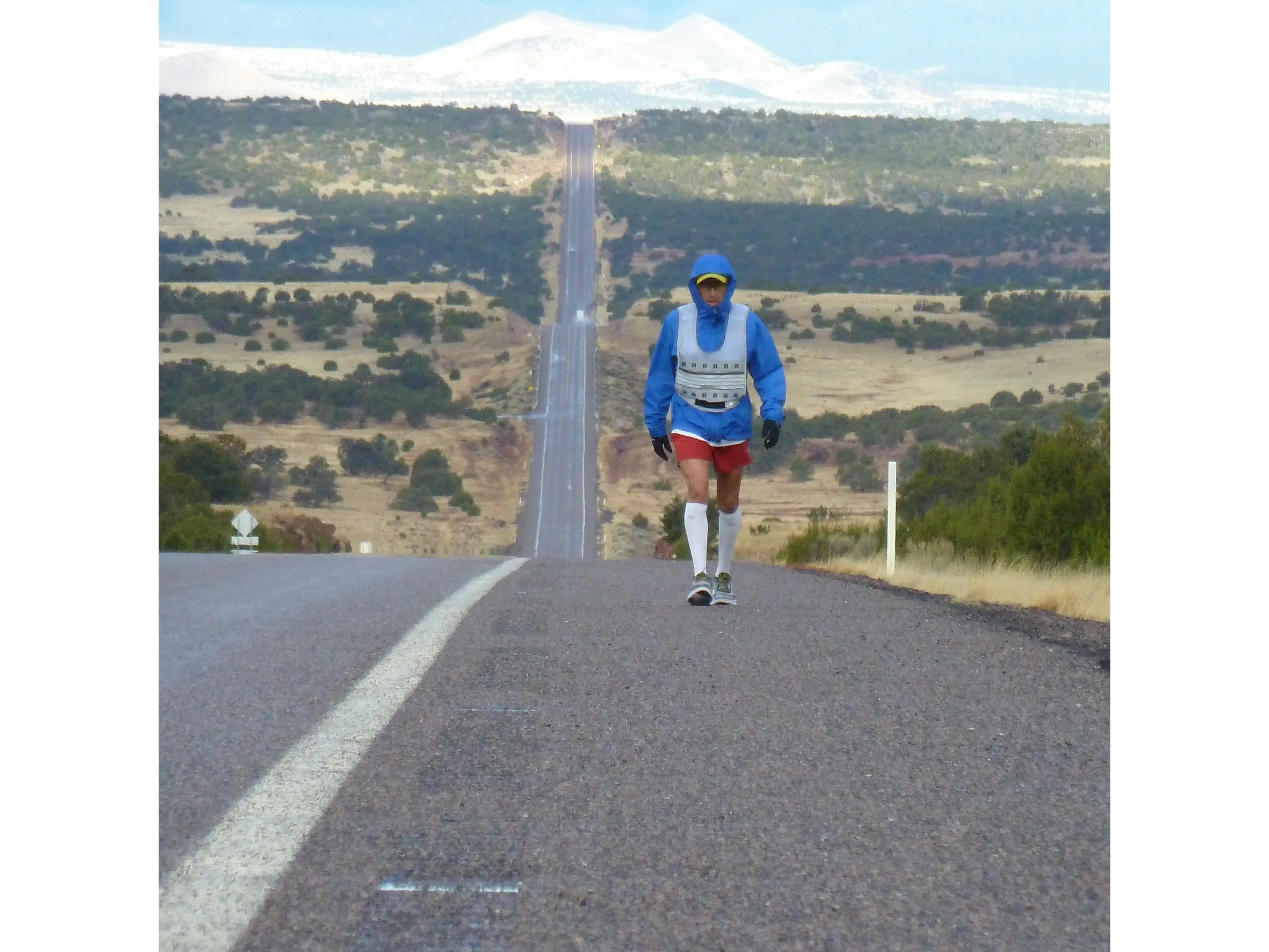A runner in blue jacket walking on a long, empty road in a mountainous landscape.