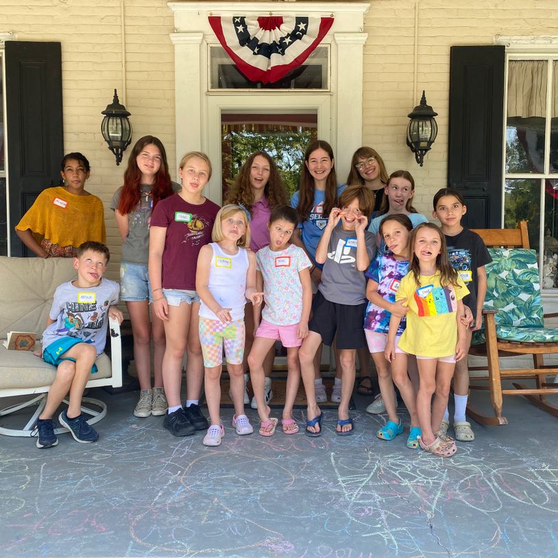 Group of kids and a woman posing playfully on a porch with chalk drawings on the ground.