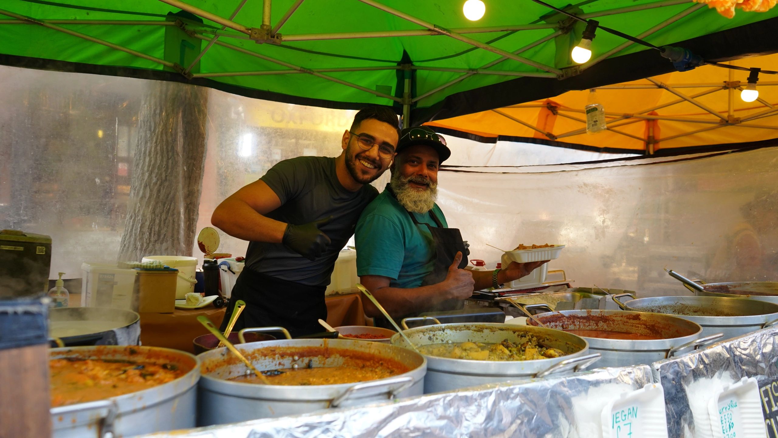 Two men smiling and giving thumbs up at a food stall with various dishes.