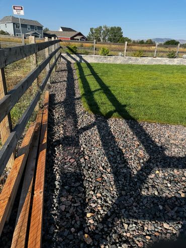 Shadows cast by a wooden fence on gravel next to a green lawn.