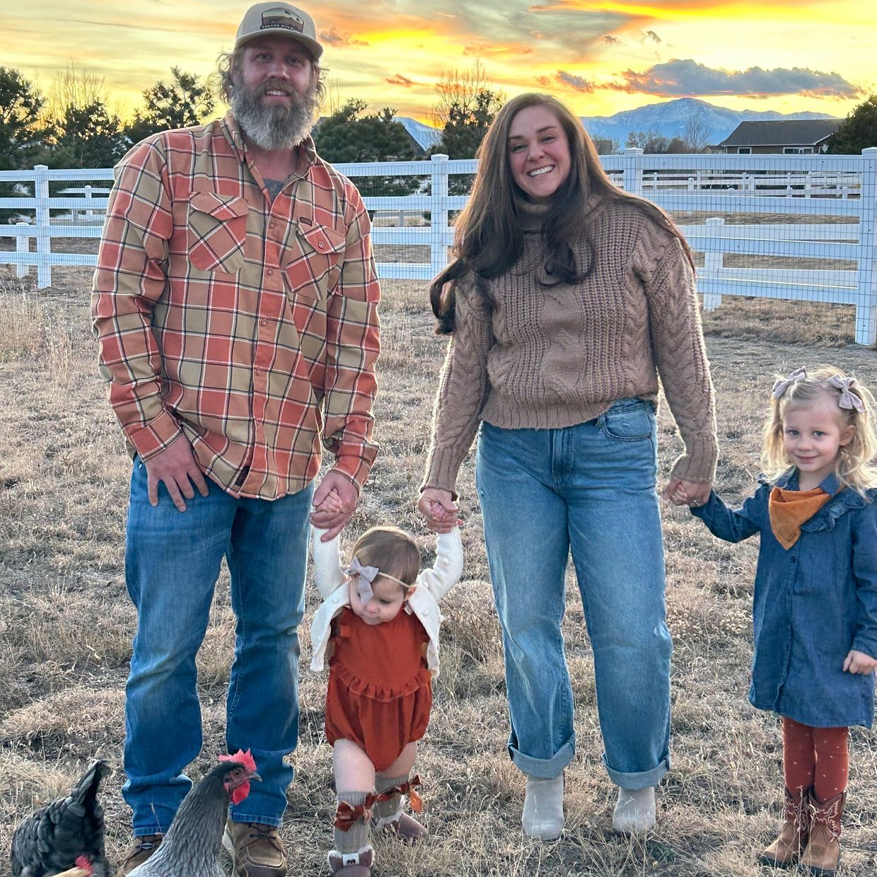 A family of four holding hands outdoors with chickens and a sunset in the background.