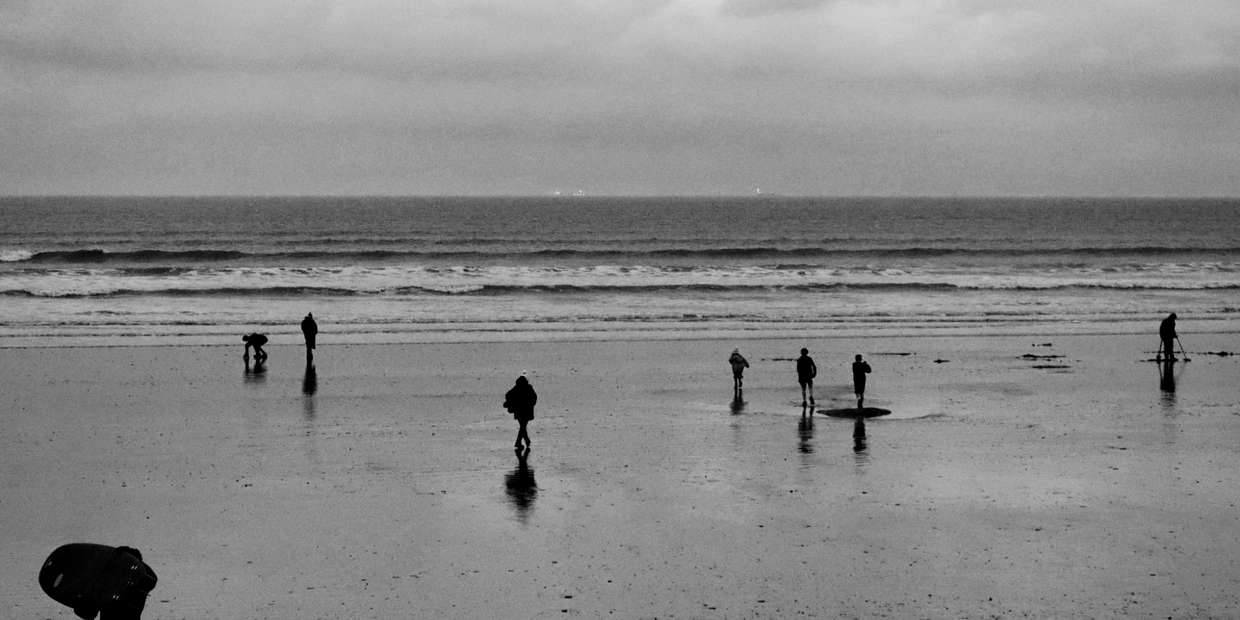 People walking and exploring a wet beach under a cloudy sky in black and white.