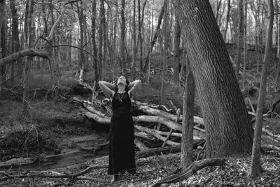 A woman in a black dress stands barefoot in a forest, looking upwards.