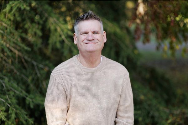 Man in a beige sweater smiling outdoors with greenery in the background.