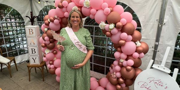 Pregnant woman smiling at her baby shower with pink balloon arch.