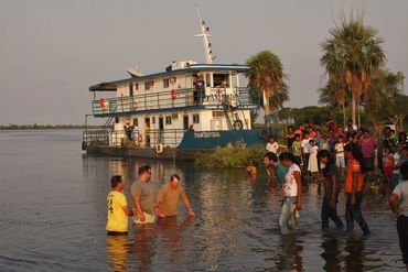 People wade through shallow water near a ferry with a crowd watching from the shore.