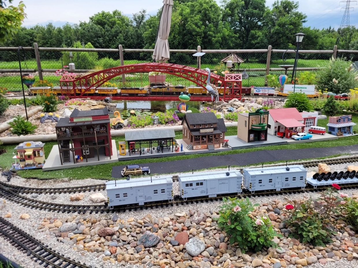 Detailed miniature train set with buildings and a red bridge in a garden.