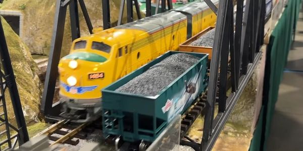 Model yellow train crossing a black metal bridge with coal cars.