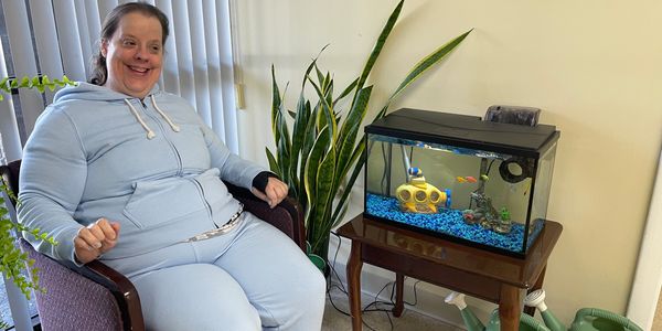 A woman in a light blue hoodie sitting next to a fish tank with colorful fish.