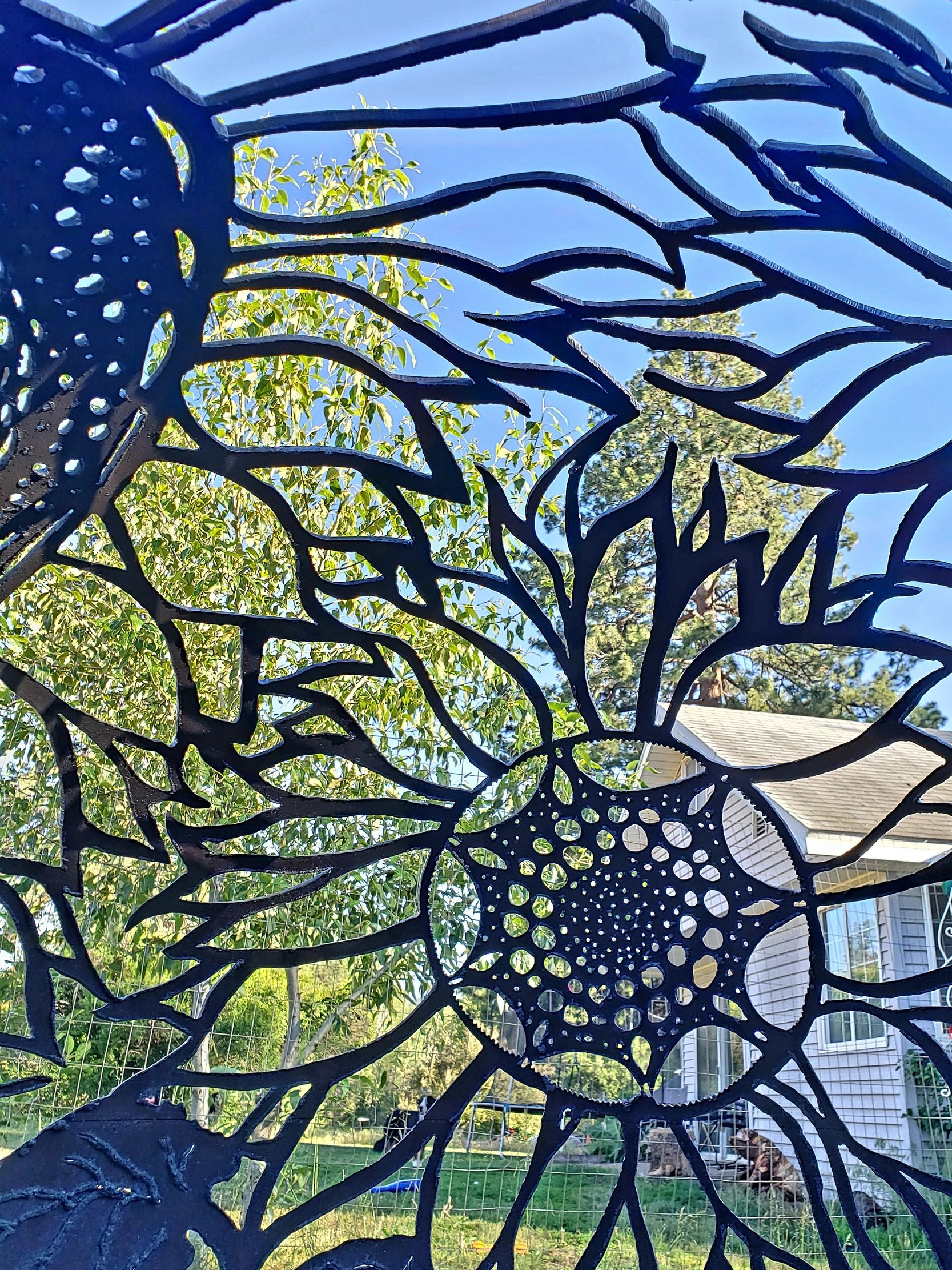 Close-up of black metal sunflower cutout against a house and trees.