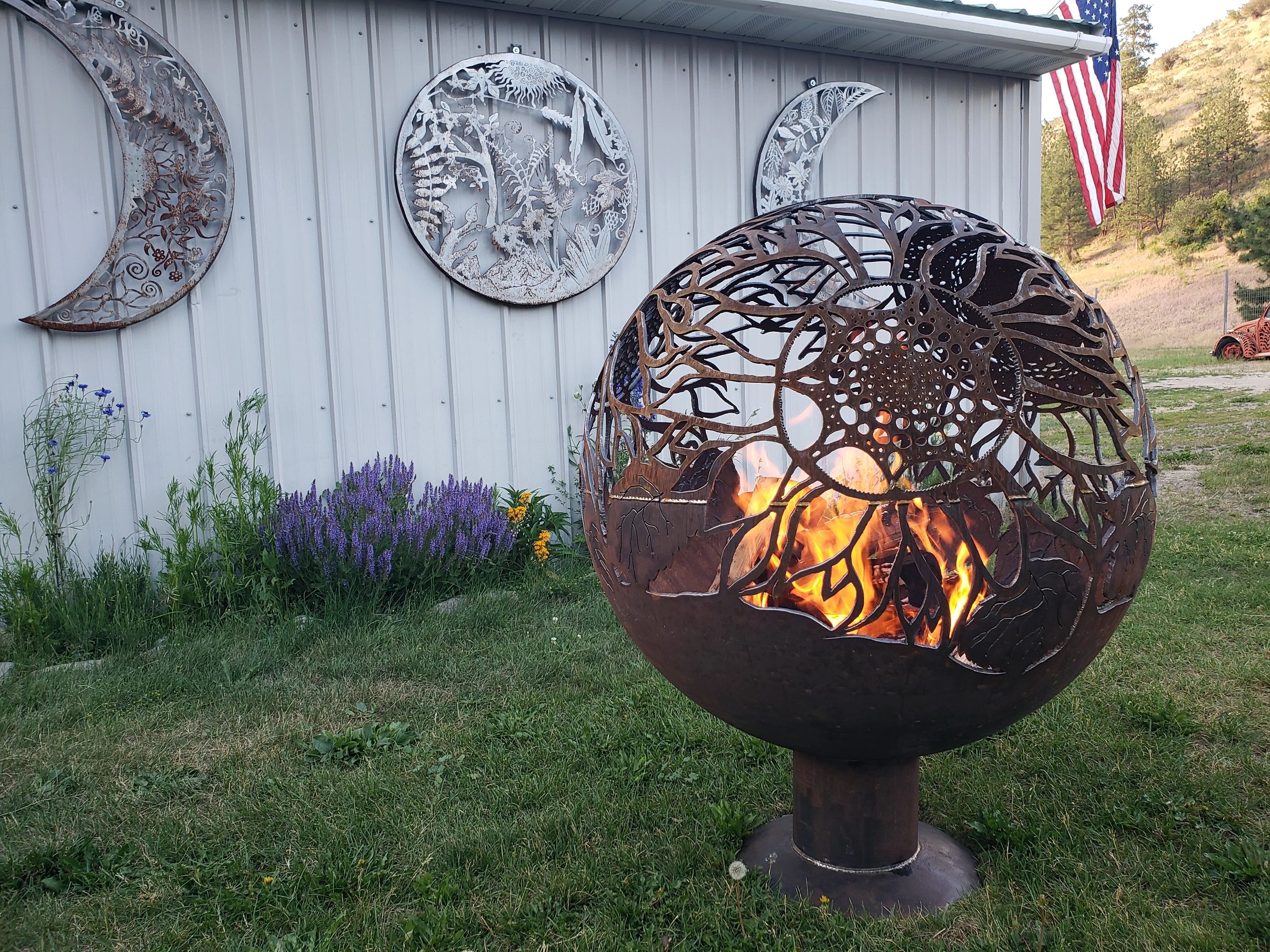 Decorative fire pit and metal art on a building exterior with flowers and an American flag.