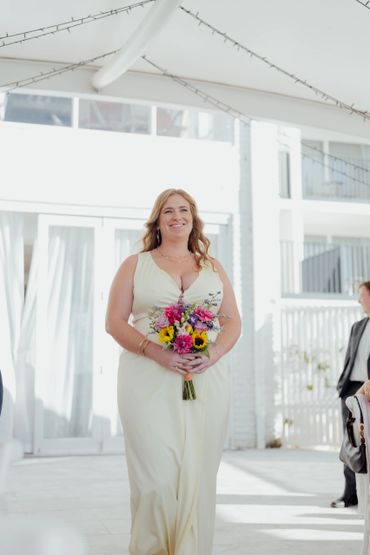 Smiling woman in a pale yellow dress holding a colorful bouquet indoors.