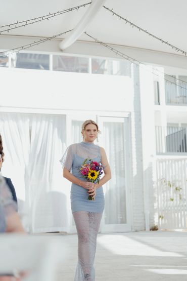 Woman in a blue lace dress holding a colorful bouquet at a bright event.