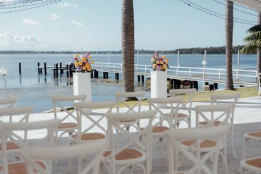 Outdoor wedding setup by the water with floral arrangements and white chairs.