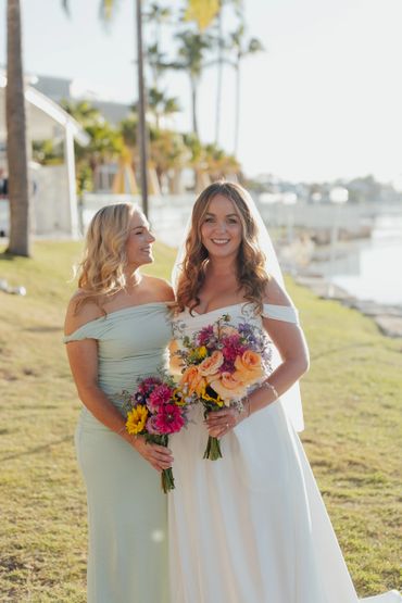 Bride and bridesmaid holding colorful bouquets outdoors on a sunny day.