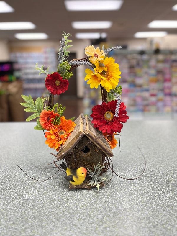 Decorative birdhouse with vibrant flowers and a small yellow bird on a table.