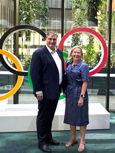 Two people posing in front of Olympic rings indoors.
