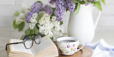 eyeglasses and an opened book on a table with coffee and a vase of flowers