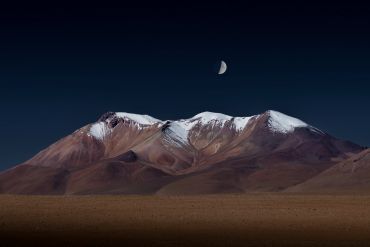 Moon above snow-capped mountain peaks at night.