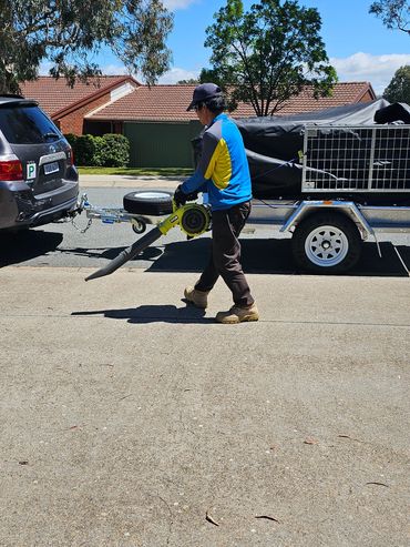 Person using a leaf blower on a sunny day near a trailer and car.