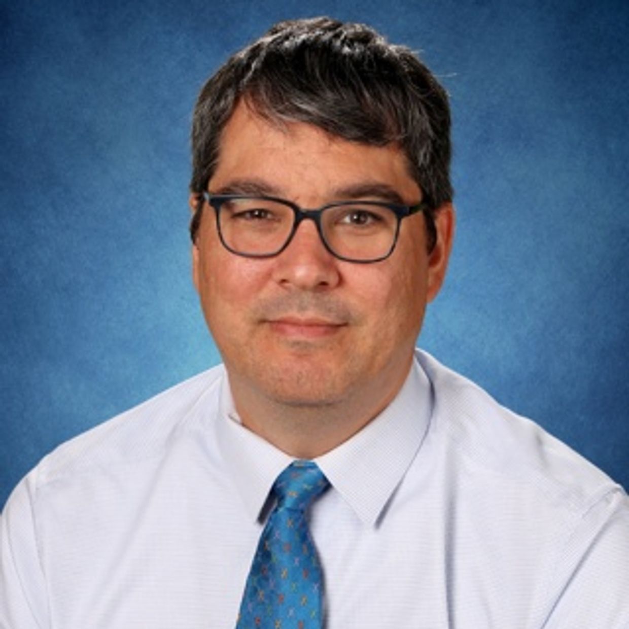 Professional man wearing glasses, white shirt, and blue tie against a blue background.