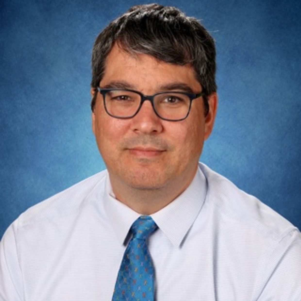 Professional man wearing glasses, white shirt, and blue tie against a blue background.