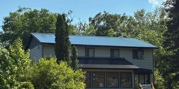 Two-story house with a blue metal roof surrounded by trees under a clear sky.