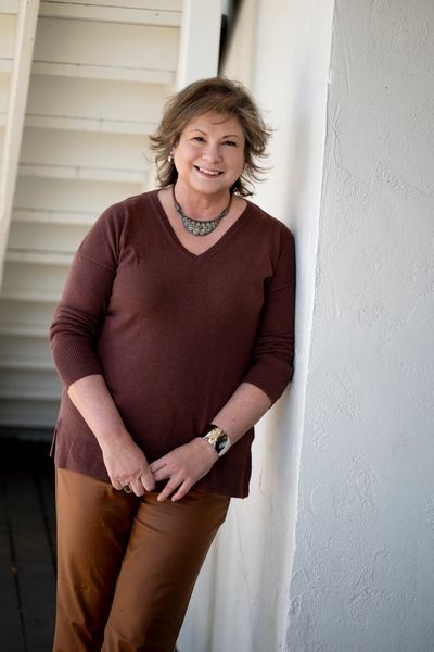 A woman in a brown sweater and pants leans against a white wall, wearing a detailed necklace