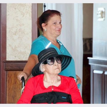 A woman in a teal shirt pushes an elderly woman in a wheelchair wearing a red top and large black hat indoors.