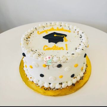 A white graduation cake decorated with black, white, and yellow dots and a graduation cap on top.