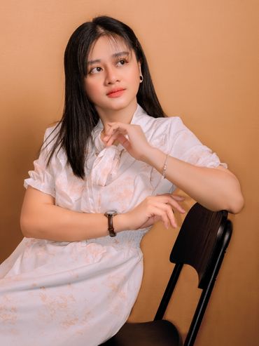 Young woman in a white dress sitting on a black chair against a tan background.