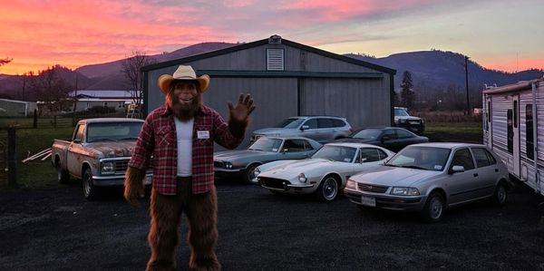 Person in Bigfoot costume waving in front of parked cars at sunset.